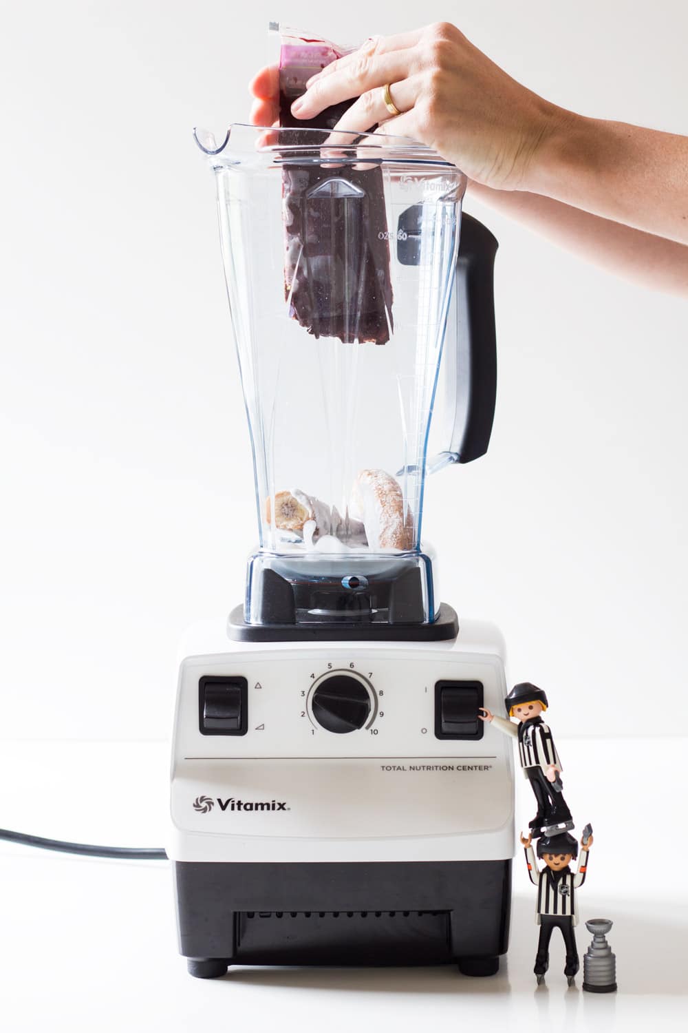 Hands squeezing acai pulp into a blender. Two toy plastic men climbing on the blender.