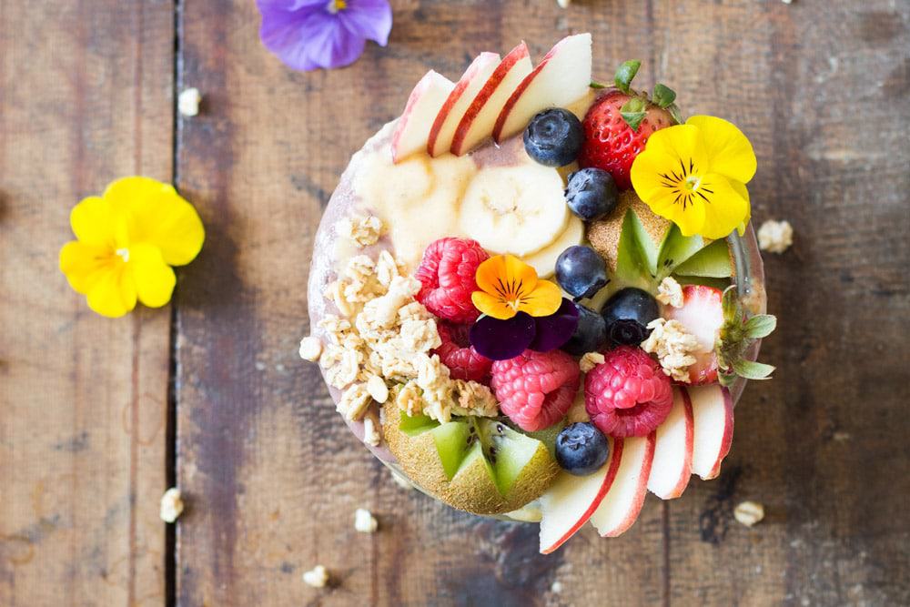 Top view of Acai Bowl topped with fresh fruit and flowers, on a rustic wood table with flowers.