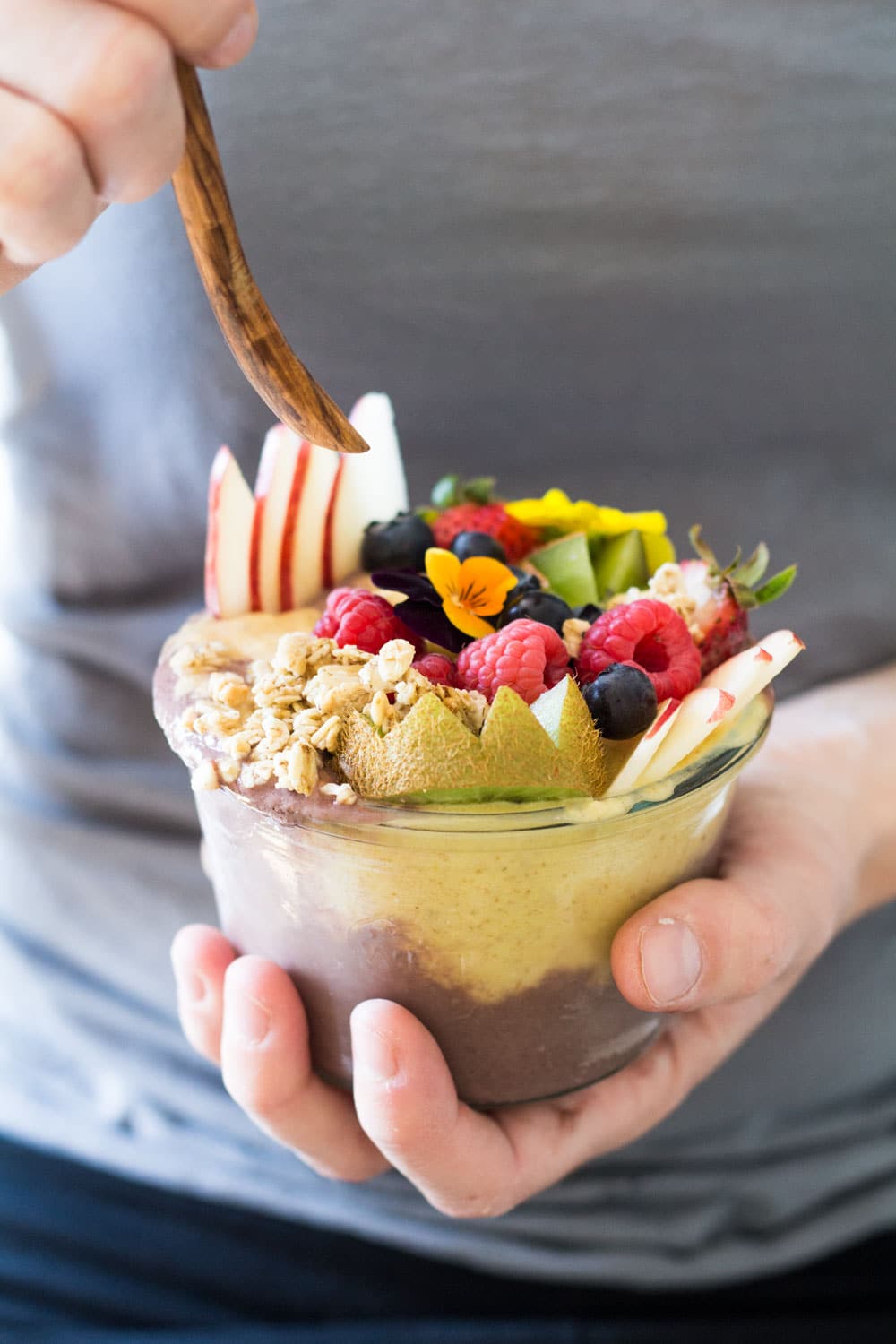 Hands holding a wooden spoon and an Acai Bowl topped with fresh fruit and flowers.