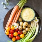 Top view of plate of fresh carrots, cherry tomatoes, green onions and baby potatoes, and a bowl of Huancaina dip.