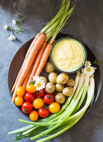 Top view of plate of fresh carrots, cherry tomatoes, green onions and baby potatoes, and a bowl of Huancaina dip.