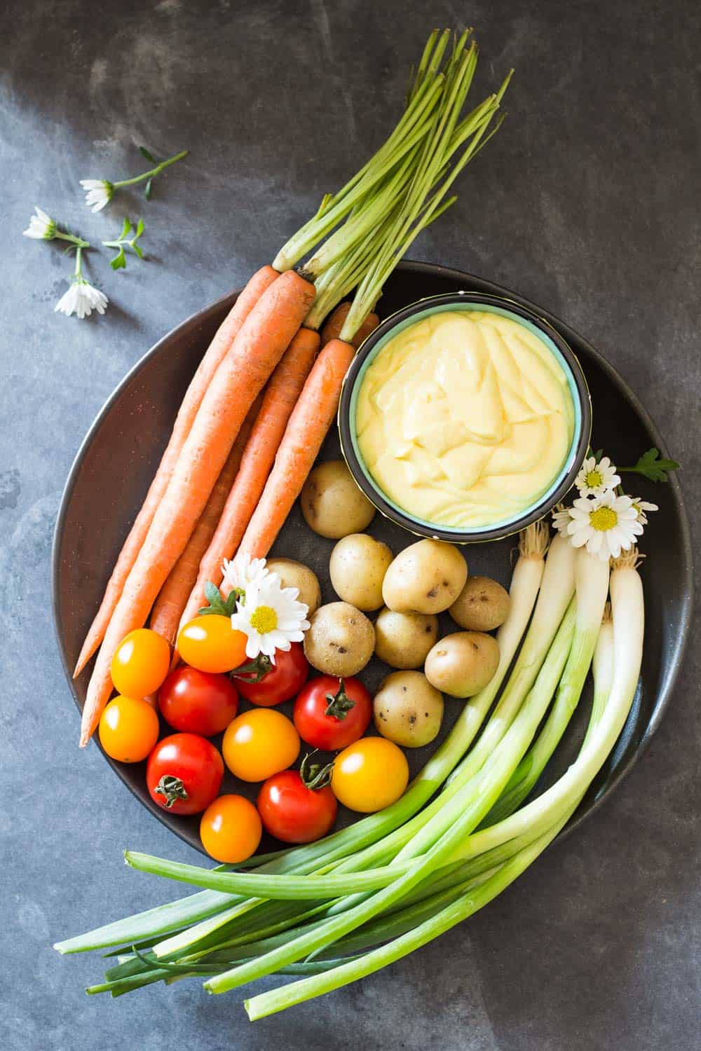 Top view of plate of fresh carrots, cherry tomatoes, green onions and baby potatoes, and a bowl of Huancaina dip.