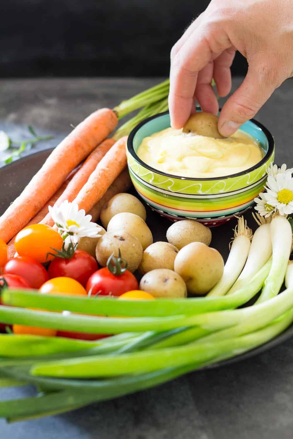 Plate of fresh carrots, cherry tomatoes, green onions and baby potatoes, and a hand dipping a potato into Huancaina dip.