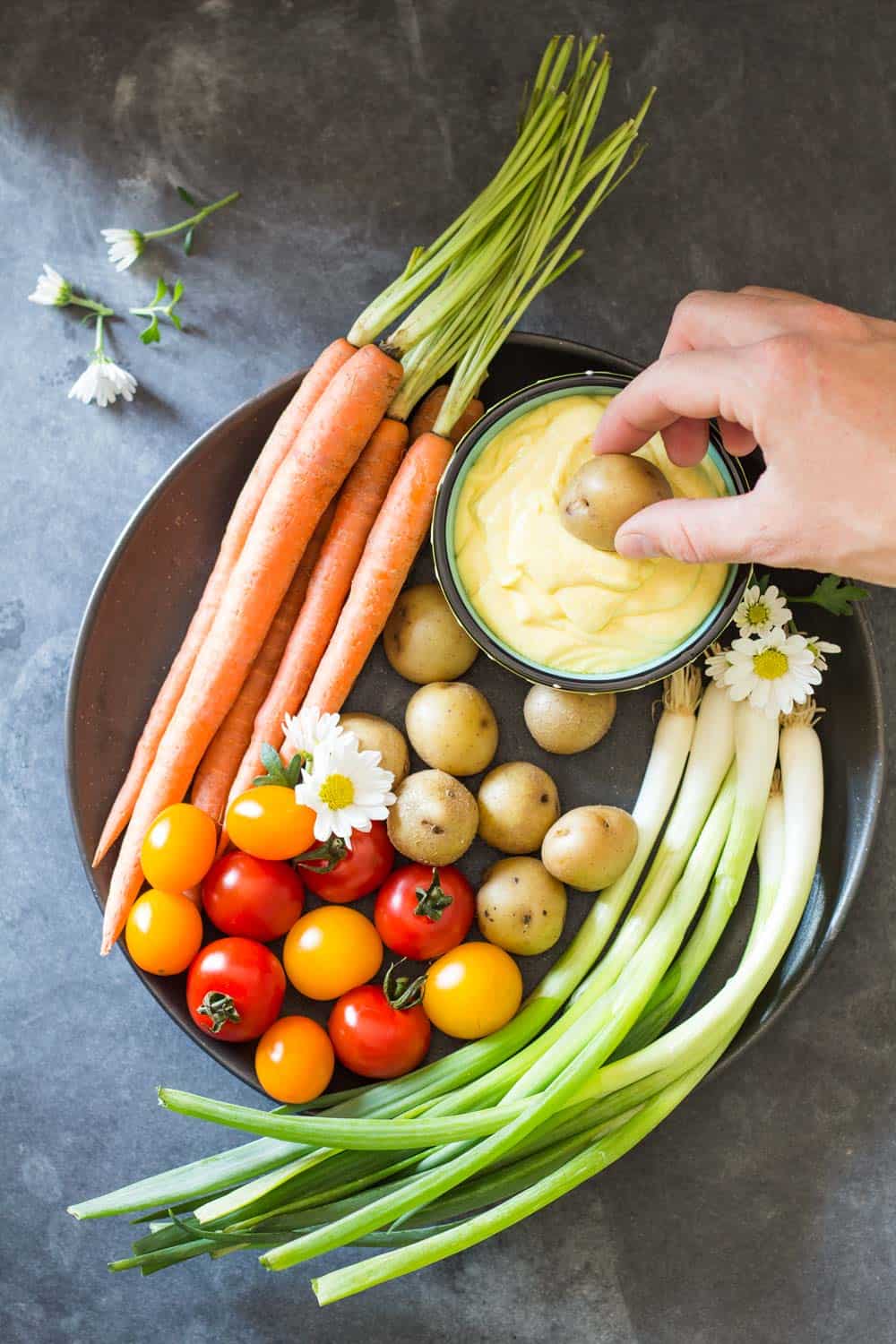 Top view of plate of fresh carrots, cherry tomatoes, green onions and baby potatoes, and a hand dipping a potato into Huancaina dip.