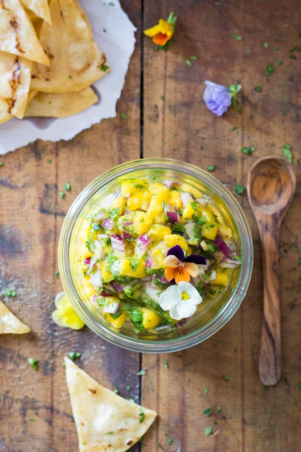 Top view of jar of Mango Salsa topped with fresh flowers, a wooden spoon, and tortilla chips.