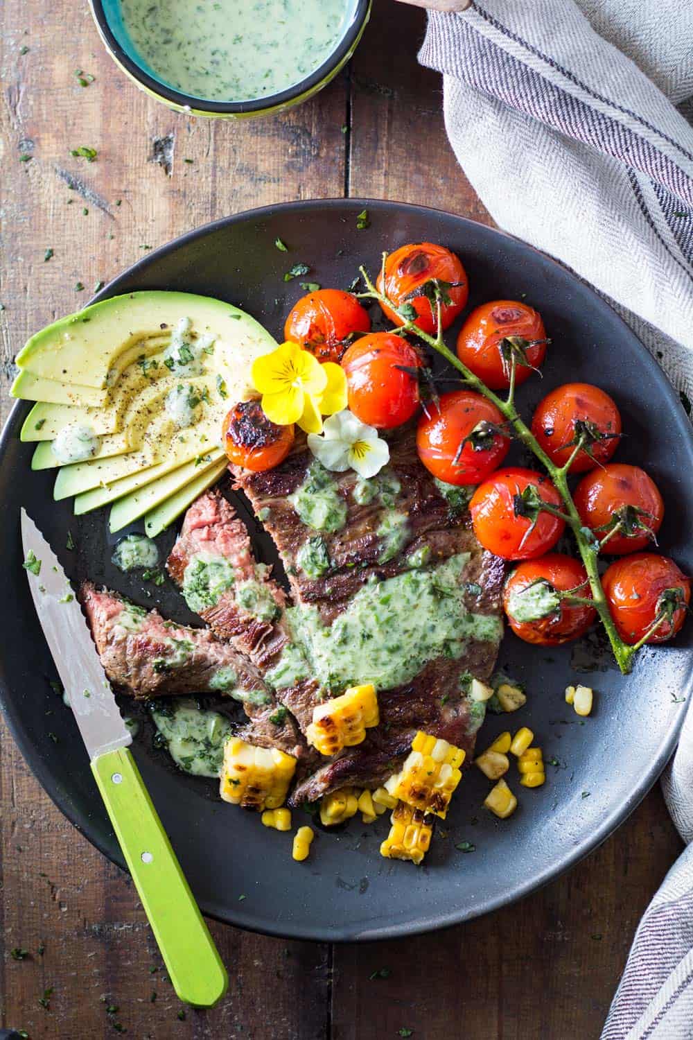 Skirt Steak with Basil Cream and Grilled Tomatoes, corn kernels and sliced avocado, a knife, and a jar of Basil Cream.