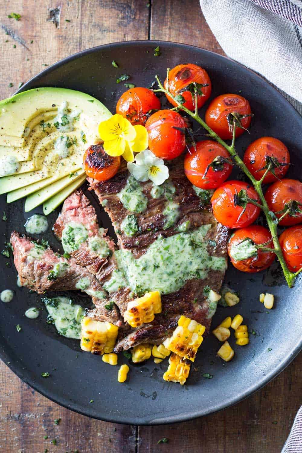 Skirt Steak with Basil Cream and Grilled Tomatoes, corn kernels and sliced avocado on a black plate.