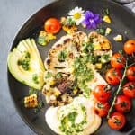 Top view of Grilled Cauliflower Steak with chimichurri sauce, cherry tomatoes and avocado on a black plate.