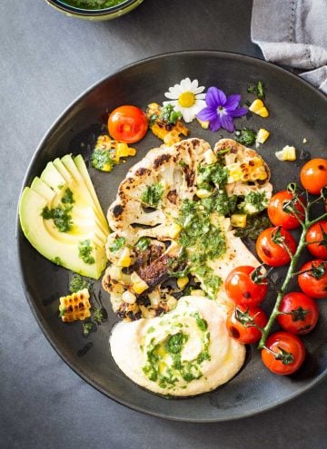 Top view of Grilled Cauliflower Steak with chimichurri sauce, cherry tomatoes and avocado on a black plate.