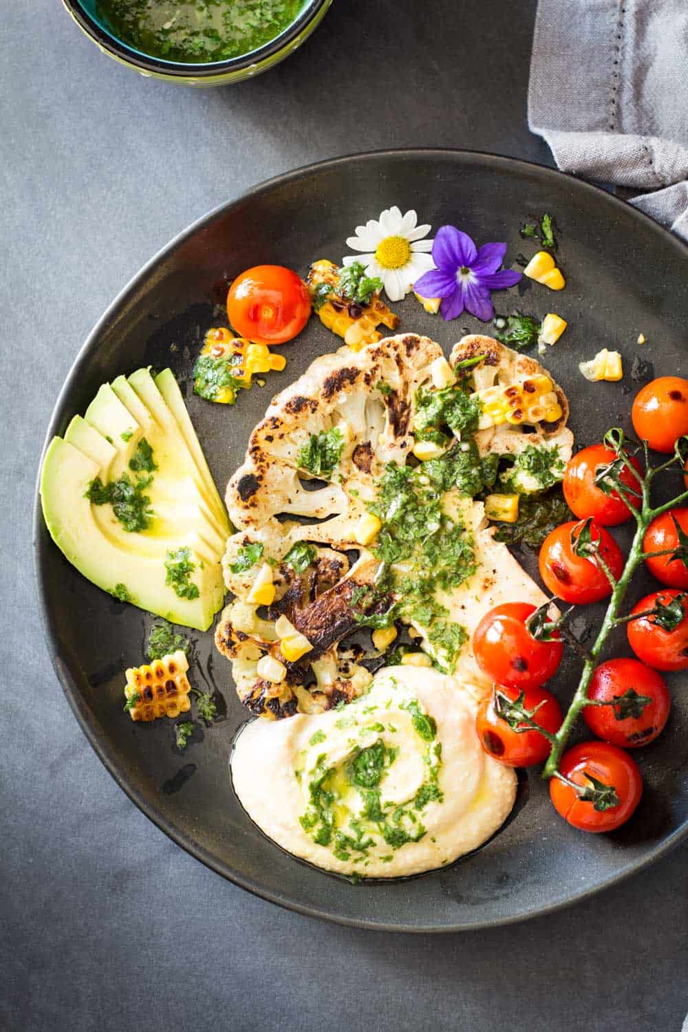 Top view of Grilled Cauliflower Steak with chimichurri sauce, cherry tomatoes and avocado on a black plate.