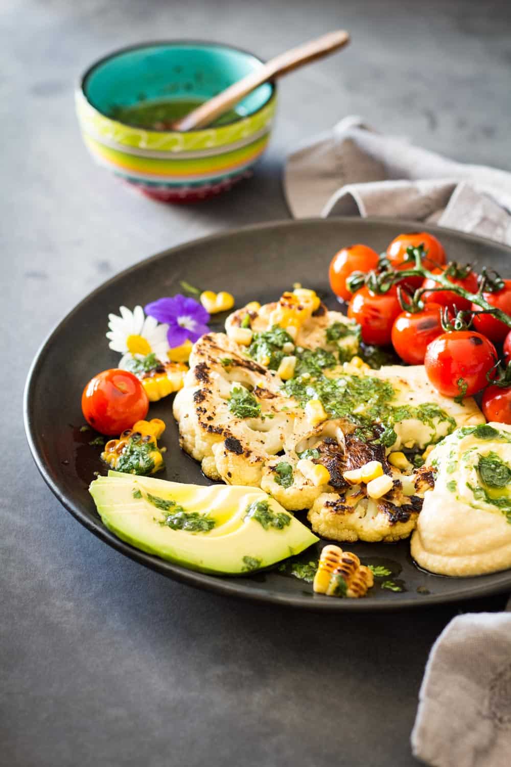 Grilled Cauliflower Steak with chimichurri sauce, cherry tomatoes and avocado on a plate, and a bowl of chimichurri sauce in the back.