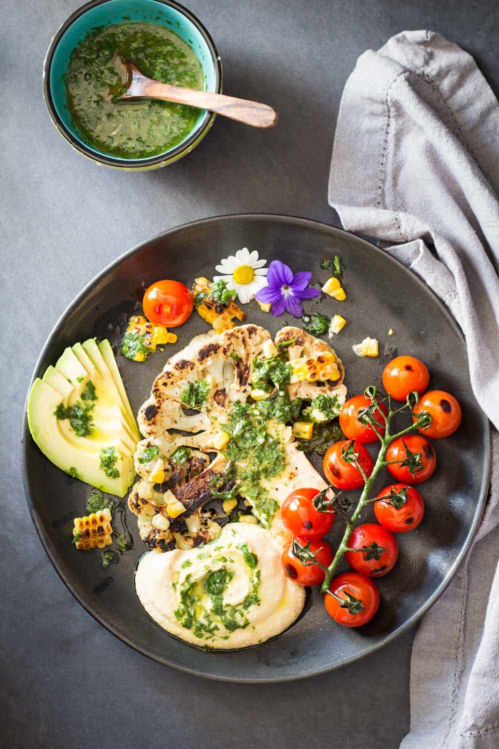 Top view of Grilled Cauliflower Steak with chimichurri sauce, cherry tomatoes and avocado on a plate, and a bowl of chimichurri sauce.