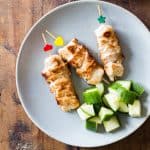 Top view of three Middle-Eastern Grilled Chicken skewers on a plate with cubed cucumber, and a small bowl of garlic sauce.