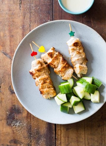 Top view of three Middle-Eastern Grilled Chicken skewers on a plate with cubed cucumber, and a small bowl of garlic sauce.
