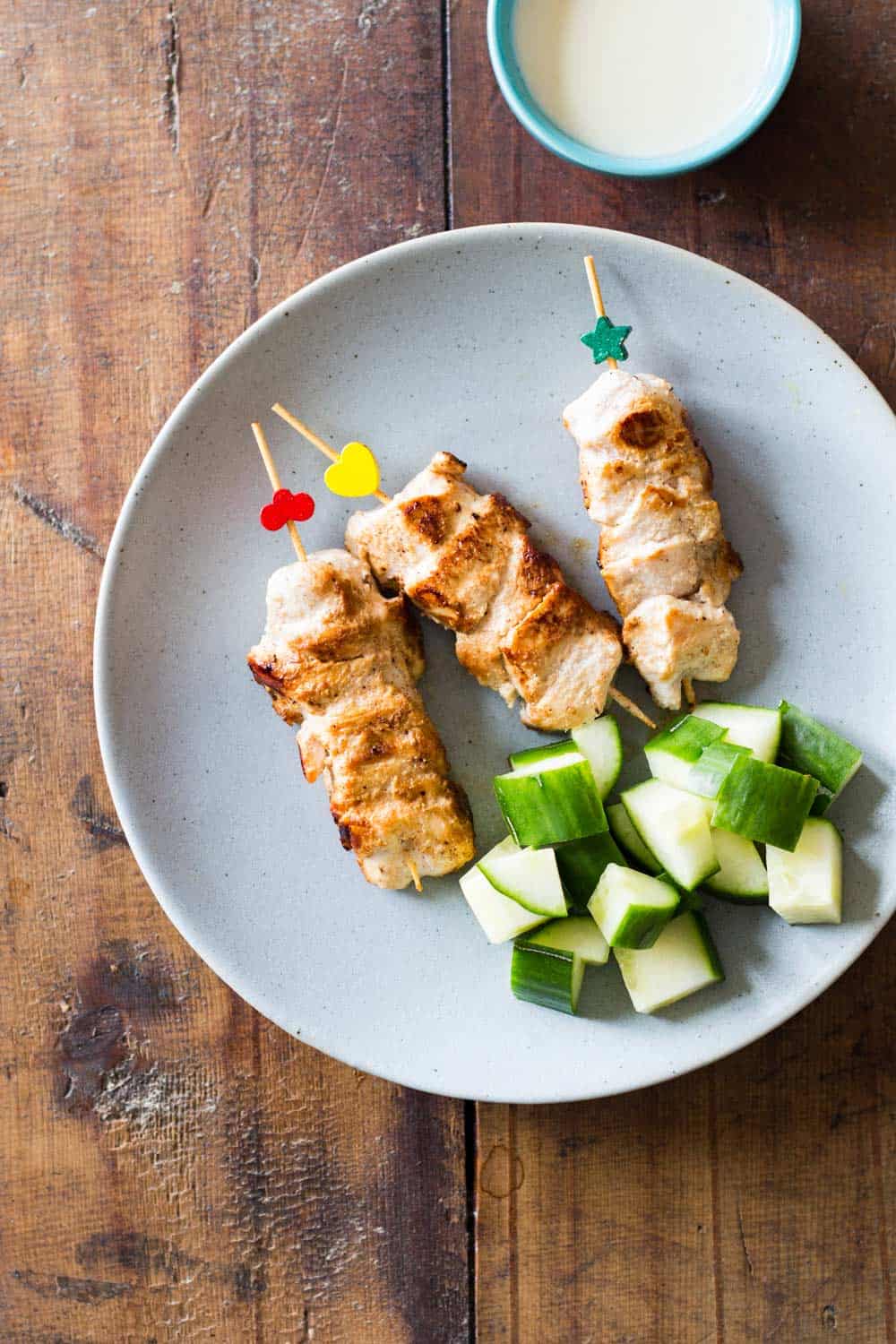 Top view of three Middle-Eastern Grilled Chicken skewers on a plate with cubed cucumber, and a small bowl of garlic sauce.