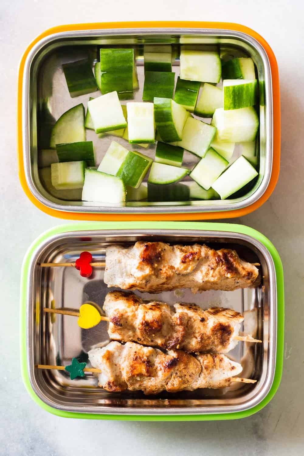 A lunch container with cubed cucumber, and another container with three Middle-Eastern Grilled Chicken skewers.