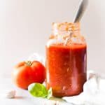 Roasted Garlic Tomato Sauce in a jar with a spoon, a fresh tomato, garlic cloves, and fresh basil leaves.