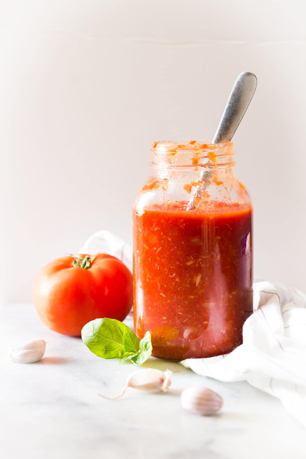 Roasted Garlic Tomato Sauce in a jar with a spoon, a fresh tomato, garlic cloves, and fresh basil leaves.