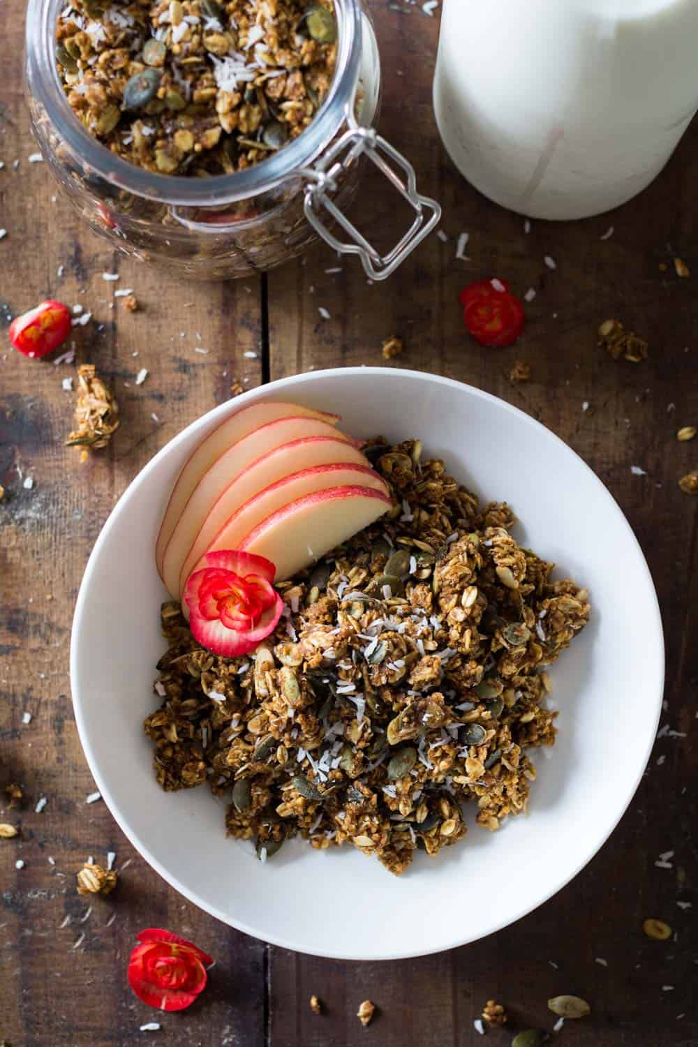 Bowl of Healthy Pumpkin Granola with apple slices, a jar with pumpkin granola, and part of a milk bottle.