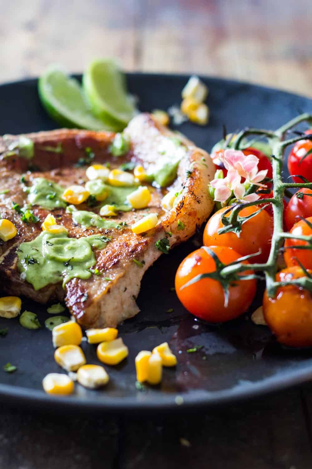 Close up of Pork Chops garnished with avocado cilantro sauce, corn kernels, and cherry tomatoes, to show texture.