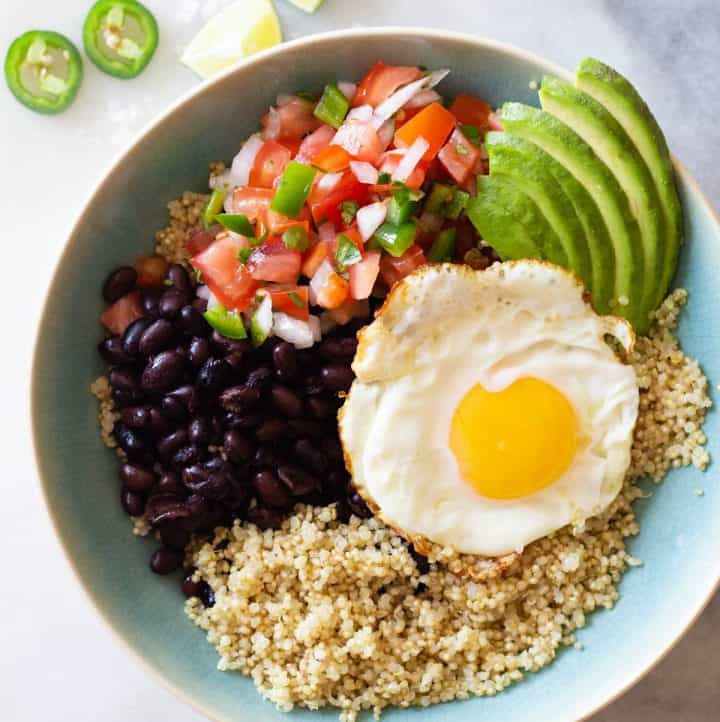 quinoa, black beans, fried egg, pico de gallo, sliced avocado in a blue bowl.