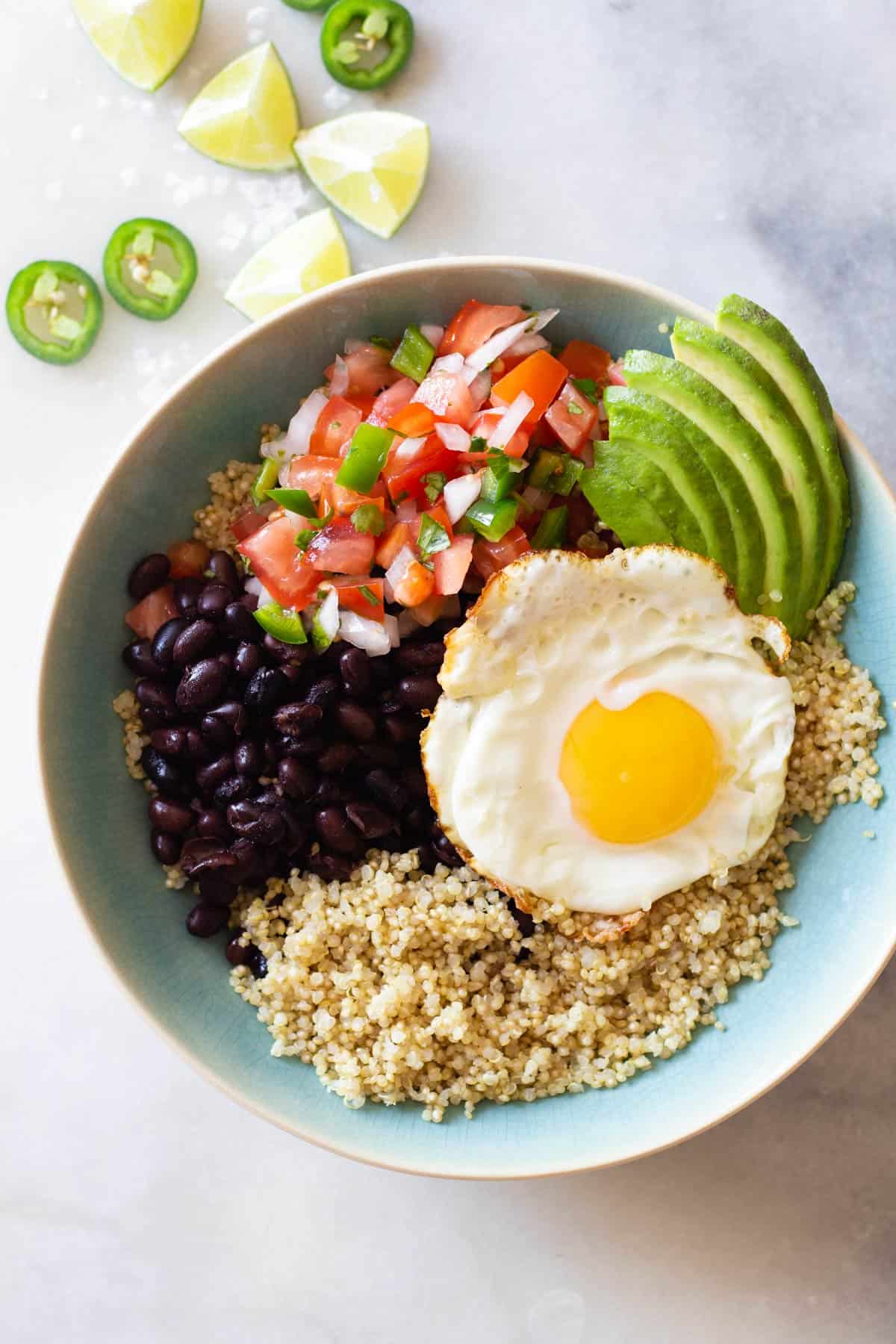 quinoa, black beans, fried egg, pico de gallo, sliced avocado in a blue bowl.