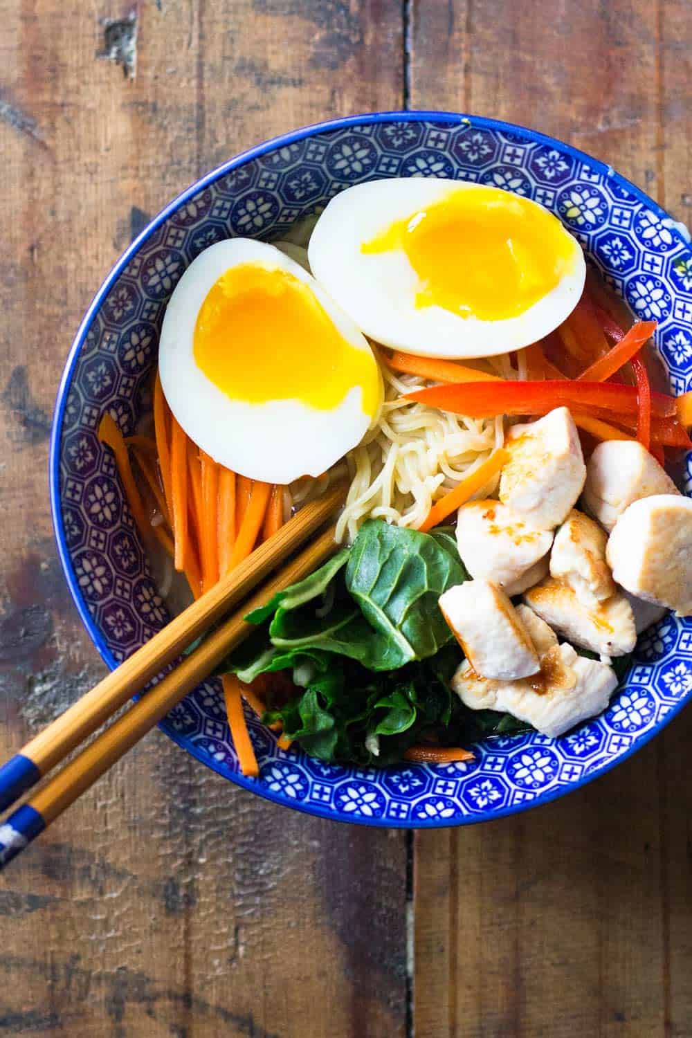 Ramen chicken noodle soup with boiled egg cut in half, served in a blue bowl with chopsticks.