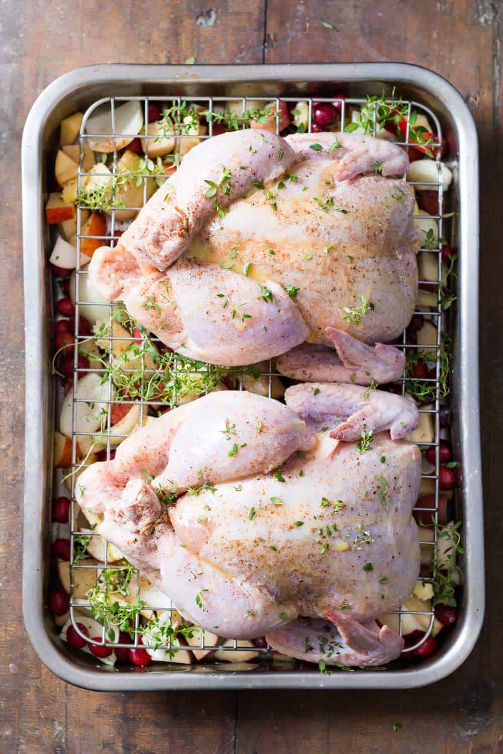 Two raw whole chickens marinated on grilling pan, with chopped apples, potatoes and cranberries at the bottom. 