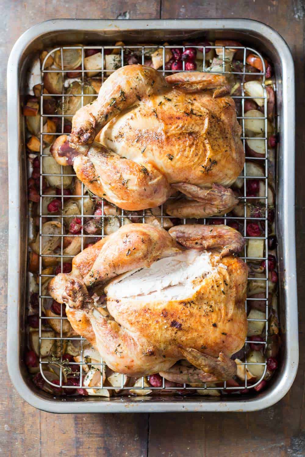 Two whole roasted chickens in grilling pan, with chopped apples, potatoes and cranberries at the bottom. 