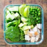 Green Green Bowl Salad with spinach leaves, cubed apple, avocado, cubed chicken, peas, cilantro, and sliced limes in a glass container.