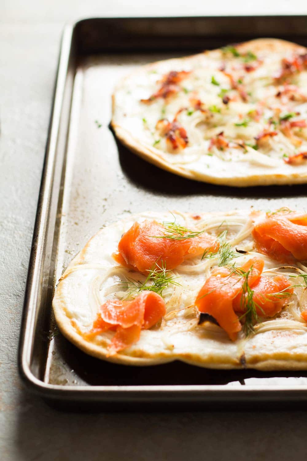 Detail of two flammkuchen. Top one with bacon and onions, bottom with smoked salmon, onions and dill, in a pan. 