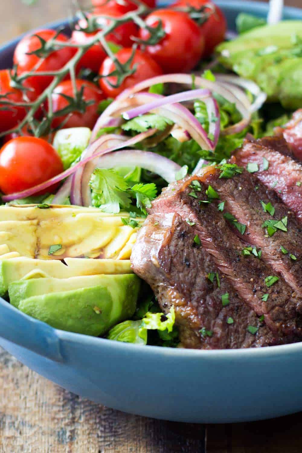 Strip loin steak salad with avocado, cherry tomatoes, cilantro and onions served in a blue bowl. 