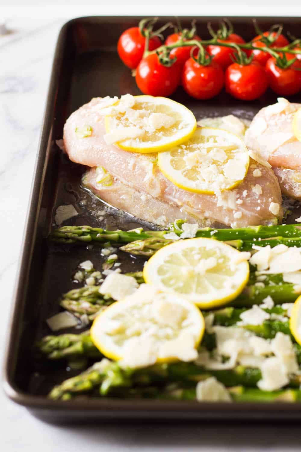 Side view of lemon chicken and asparagus, and a sprig of cherry tomatoes in a pan. 