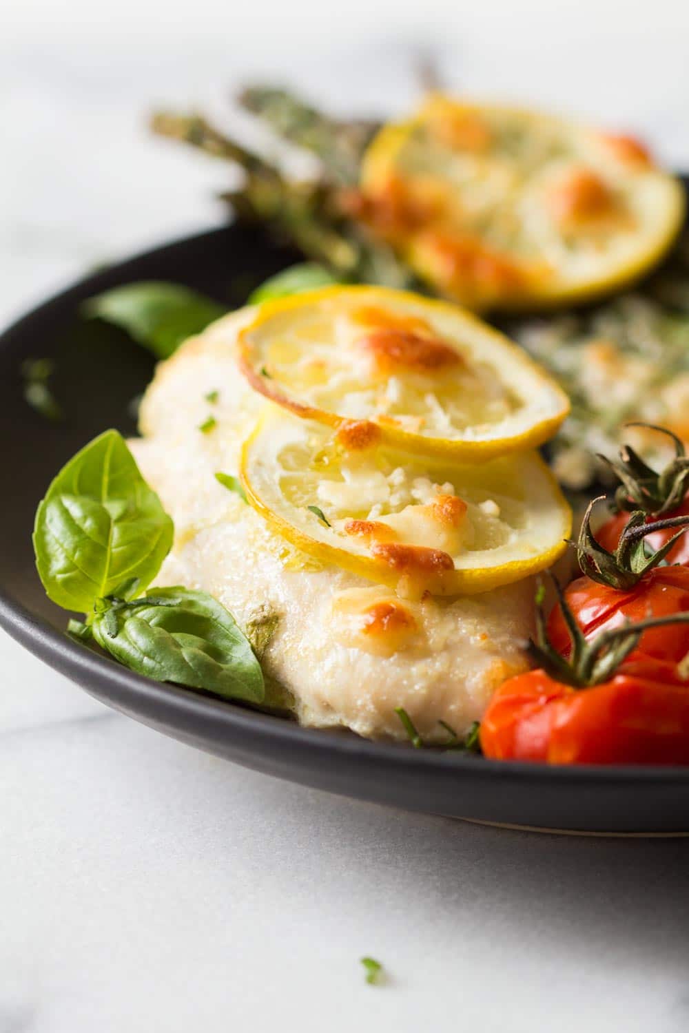 Detail of baked lemon chicken and asaparagus with cherry tomatoes, served on a black plate. 