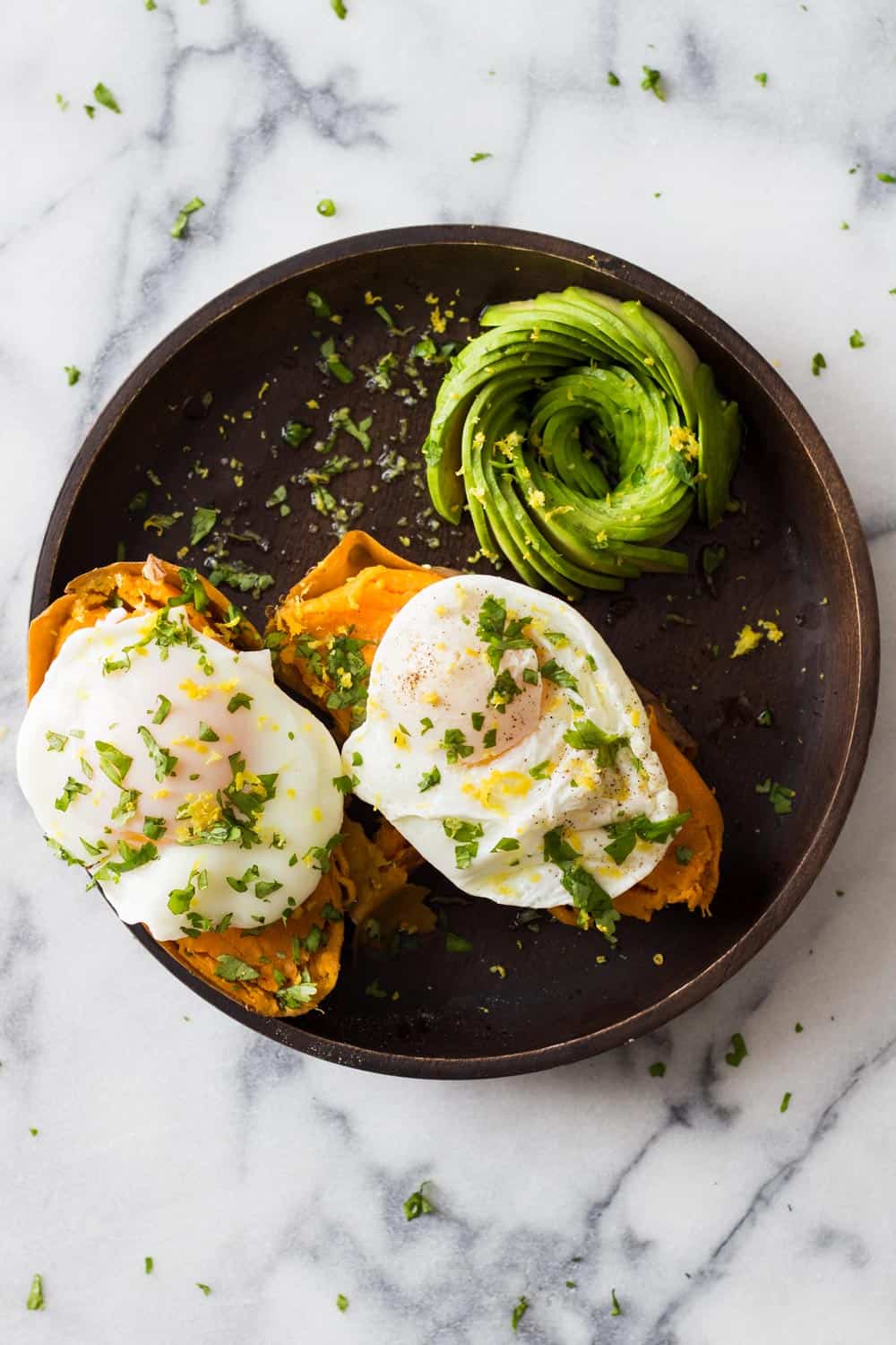 Two instant pot sweet potatoes with a poached egg on top and sliced avocado on the side, sprinkled with herbs, served on a brown plate.