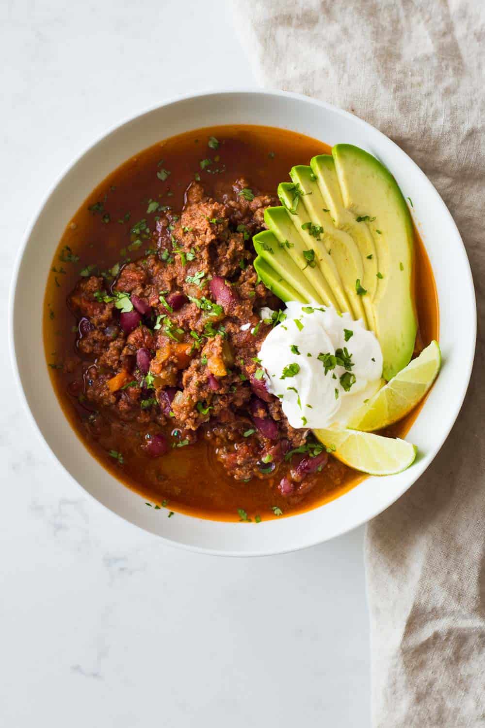 Instant Pot Chili topped with sour cream, sliced avocado and sliced lime, in a white bowl.