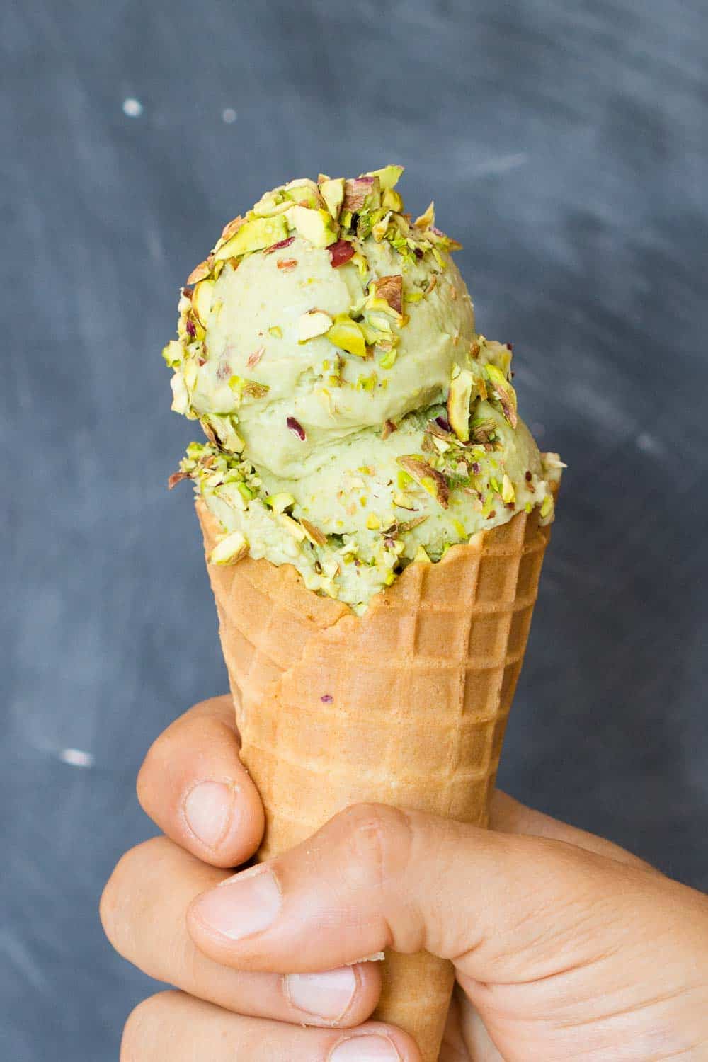Hand holding a cone with a scoop of pistachio ice cream, topped with chopped pistachio. 