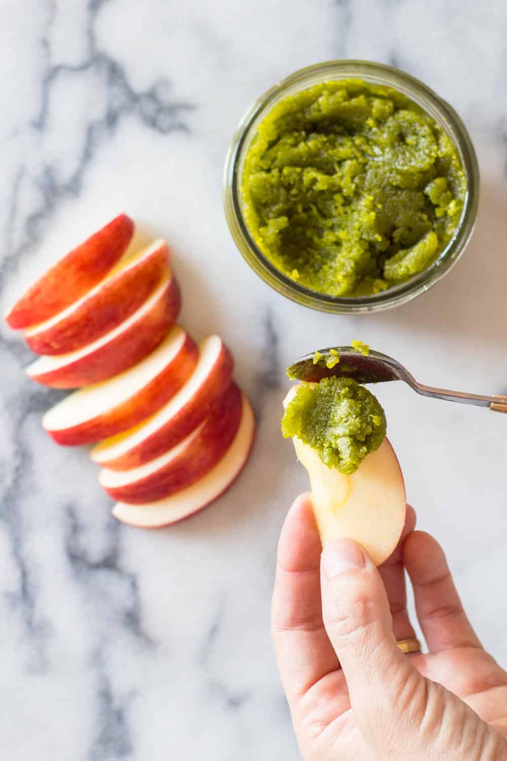 Hand holding a slice of apple being spread with pistachio paste from a jar, and some more apple slices on a marble counter top. 