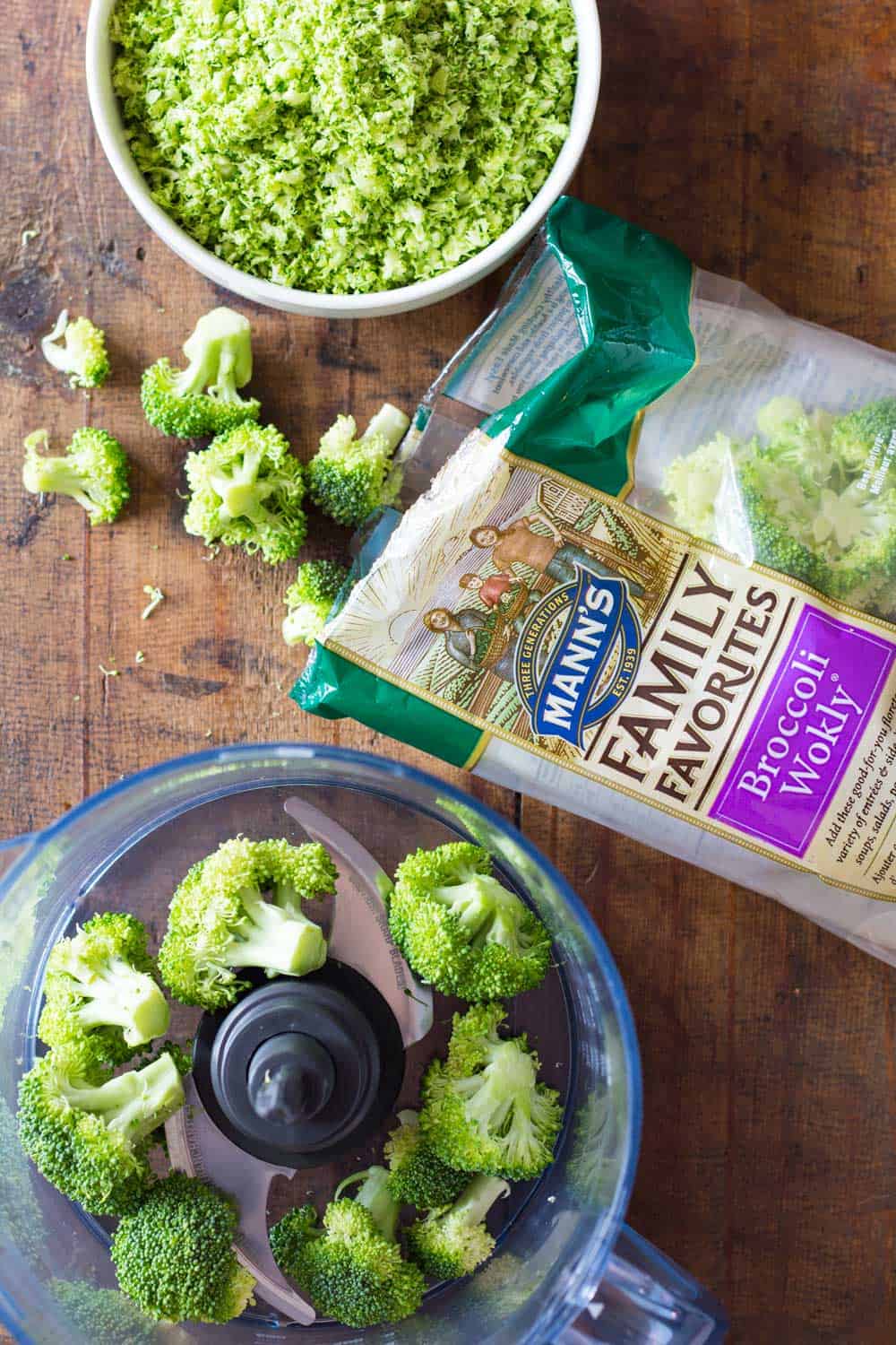 Bowl of broccoli rice, an opened bag of Mann's broccoli florets, and some broccoli florets shown inside a food processor. 