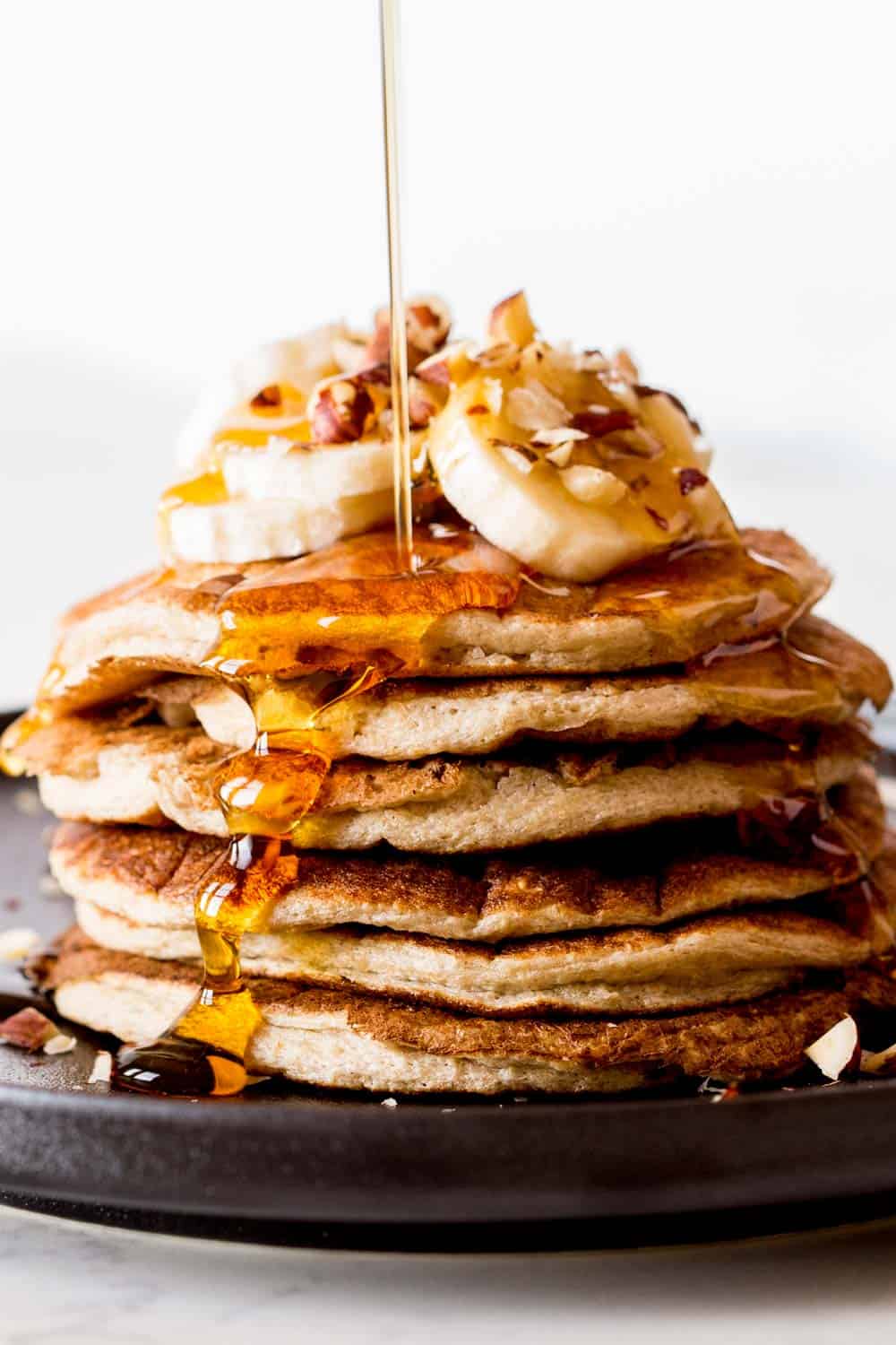 Flourless banana oatmeal pancakes stacked on a plate, topped with banana slces and chopped pecan, being poured with maple syrup