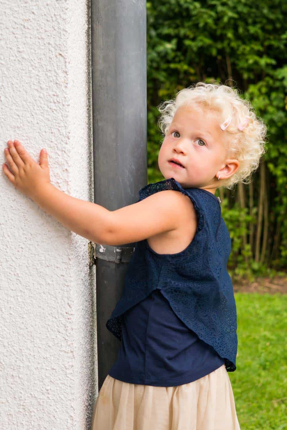 Little girl with a blue top and beige skirt hugging a wall with a post.