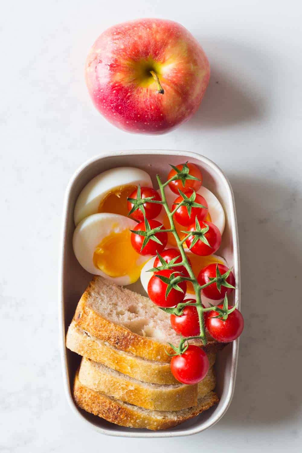 Meal prep container filled with soft boiled eggs, buttered sourdough bread, cherry tomatoes and an apple on the side.