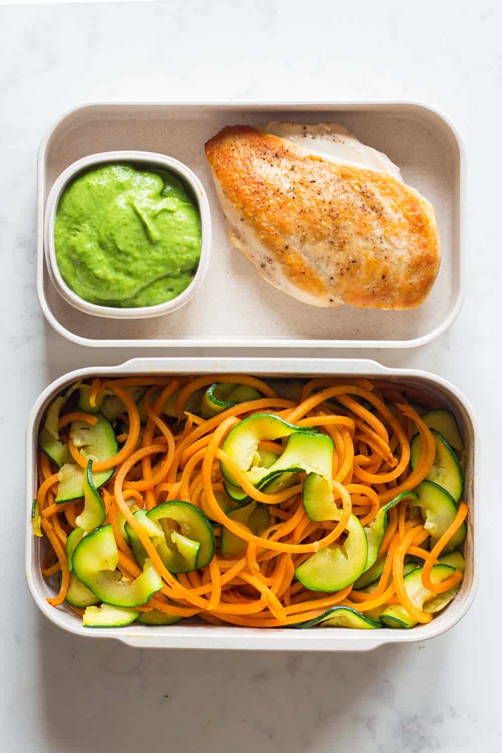 Two meal prep containers filled with pan-fried chicken breast, stir-fried spiralized sweet potato and zucchini with an avocado cilantro dressing on the side.