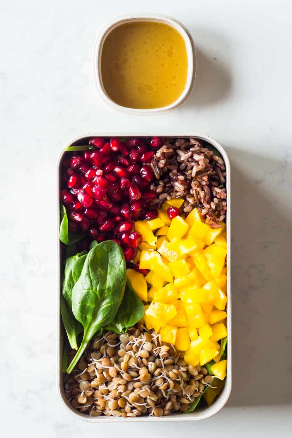 Meal prep container filled with baby spinach, cooked sprouted lentils, cooked wild rice blend, pomegranate seeds, chopped bell pepper and a homemade honey-mustard salad dressing on the side.