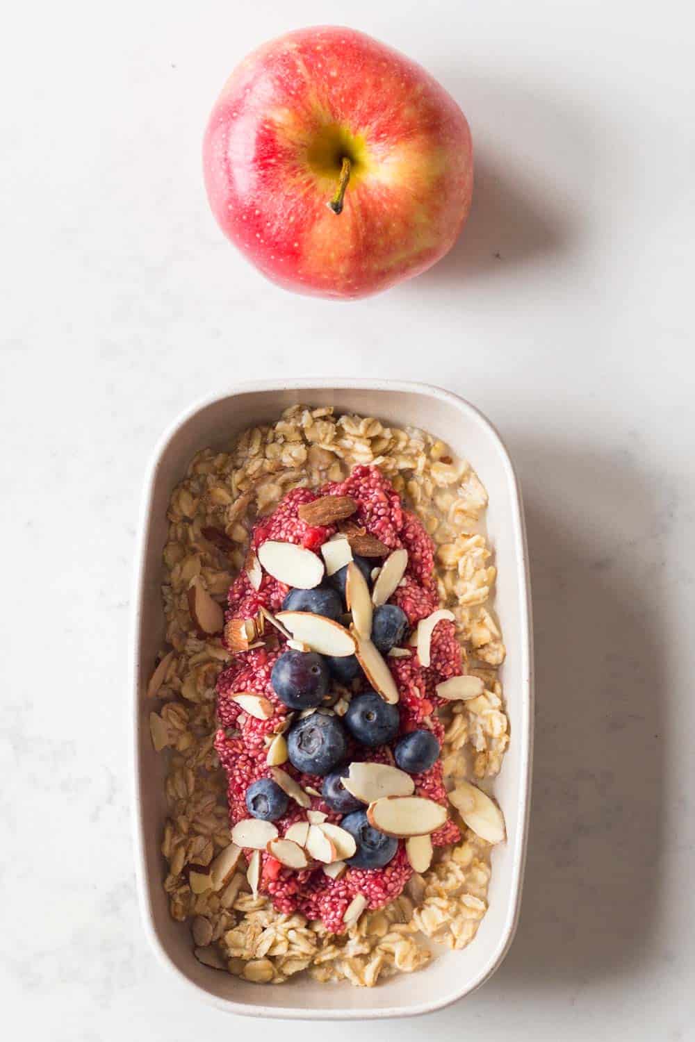 Meal prep container filled with overnight, chia pudding jam, sliced almonds, blueberries and, an apple on the side. 