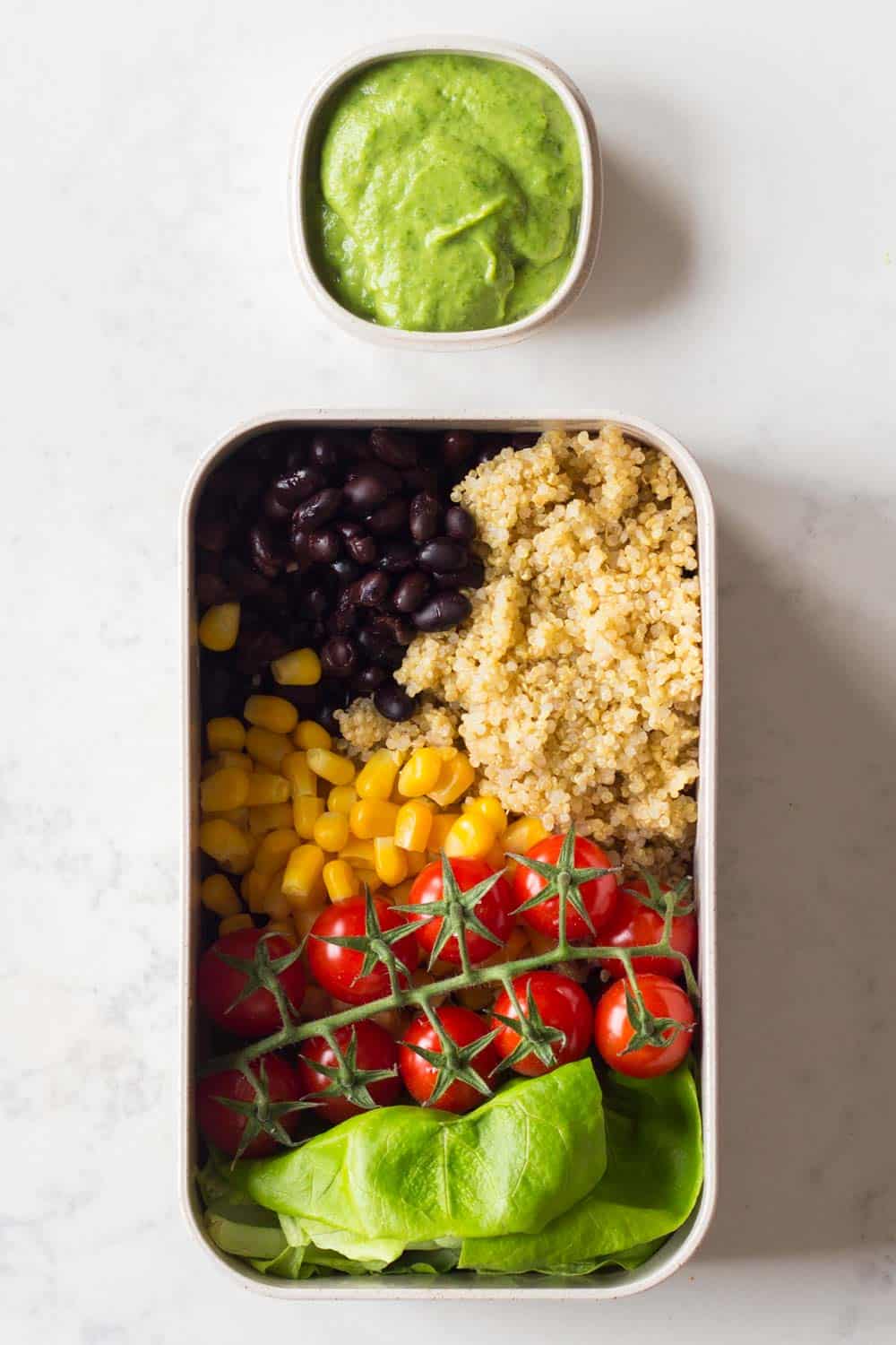 Meal prep container filled with black beans, quinoa, corn, butter lettuce and cherry tomatoes with an avocado cilantro dressing on the side.