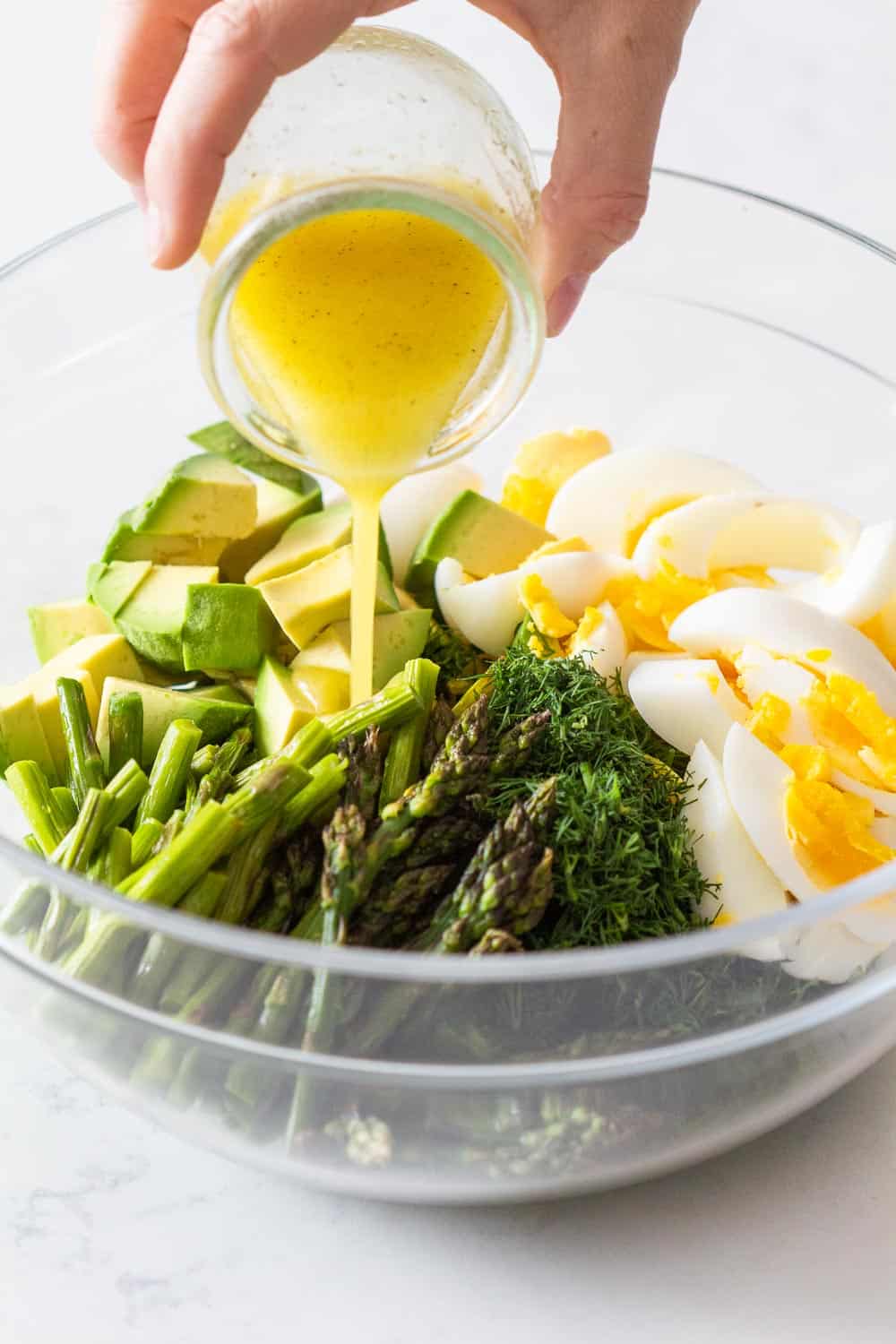 Pouring Lemon Olive Oil Dressing over Avocado and Egg Salad ingredients in glass bowl.