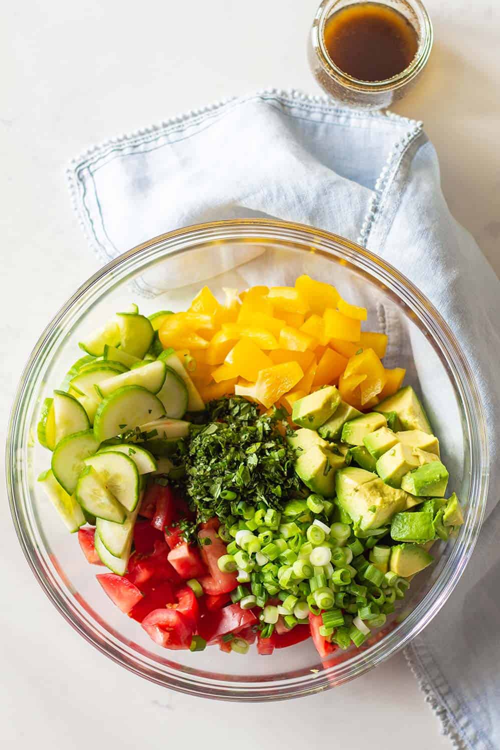 Cucumber Tomato Avocado Salad ingredients in a large glass bowl and balsamic vinaigrette in a separate glass jar