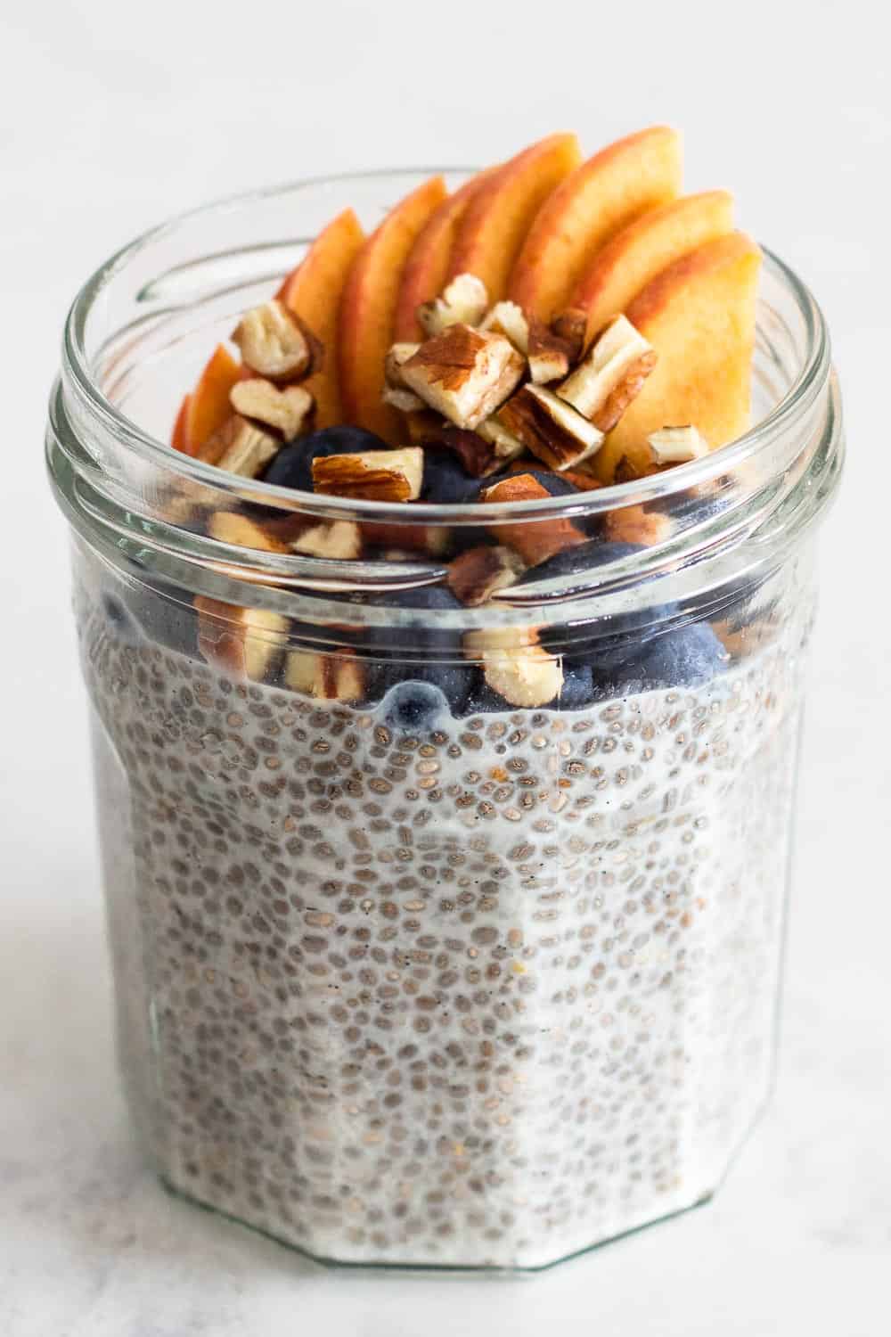 Vanilla-flavored almond milk Chia Seed Pudding in a glass jar topped with peach, blueberries and pecans.