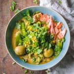 Thai Curry Soup in a blue bowl showing shredded salmon, potatoes, broccoli, and snow peas in broth on a wooden table